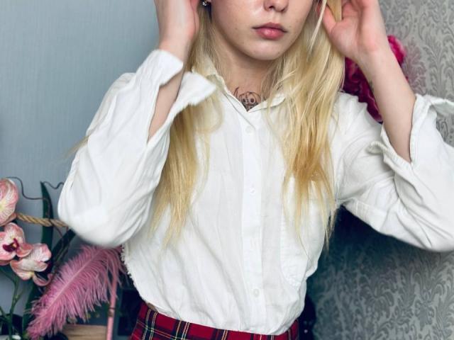 Lialit with blonde hair, white button-up shirt, and red plaid skirt, hands in hair, subtle tattoo on neck, pink flowers in background, soft pink lips, cute face.