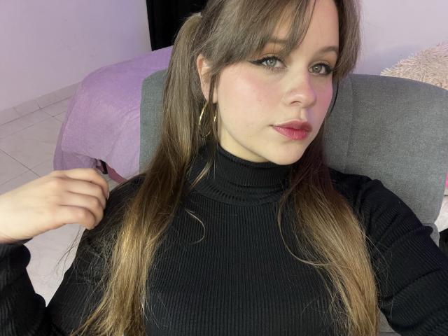 SwanKyivy met lang bruin haar in staarten, gekleed in een zwarte geribbelde coltrui, gouden oorringen en roze lippenstift, zittend op een grijze stoel. Haar hand is dichtbij haar gezicht..
