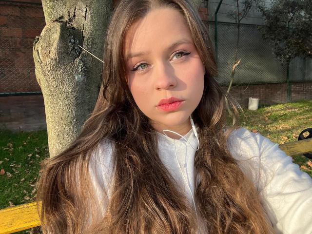 SwanKyivy, con cabello largo y castaño, camisa blanca, labios rosados y auriculares, sentada junto a un árbol, con la luz del sol en el rostro, expresión seria, un banco amarillo, césped verde y una pared de ladrillo..
