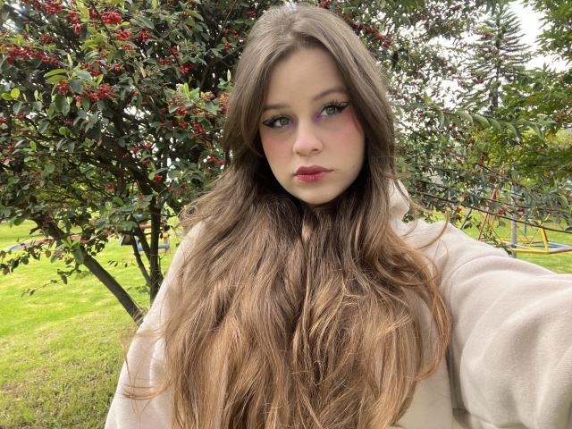 SwanKyivy, con cabello castaño largo, ojos verdes, labios rosados y un abrigo beige, se toma un selfie en un parque con césped, con un arbusto con bayas rojas detrás de ella..
