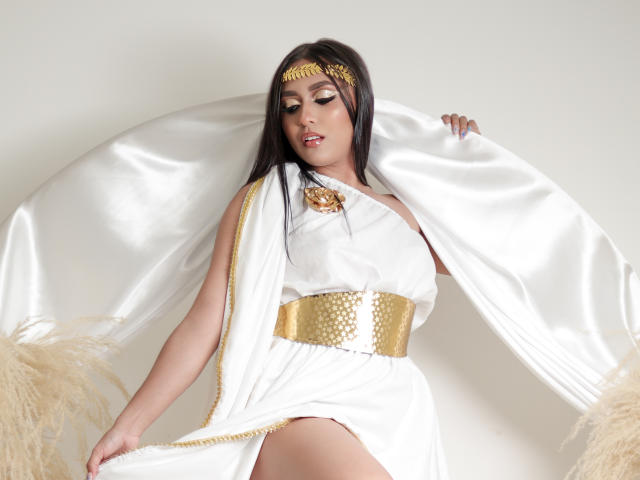SofiaJames with dark hair, white toga dress, gold headpiece, and gold belt, holding white fabric, showing her thigh and gold flower accessory. She's sexy and confident.