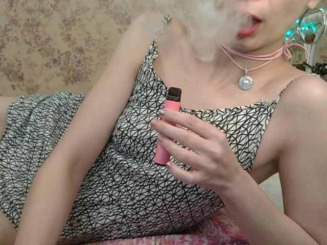 XAimeeLove with smoke in her mouth, wearing a black-and-white patterned dress, holding a pink vape, pink choker, and a silver pendant, sitting on a pink fluffy surface.