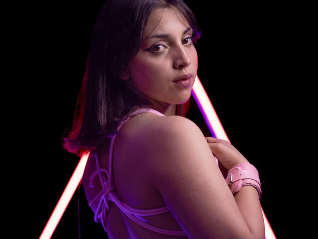 VickyButerfly with shoulder-length brown hair, pink lingerie, and pink wrist cuffs, looking over her shoulder at the camera, neon pink and purple lights in the background.