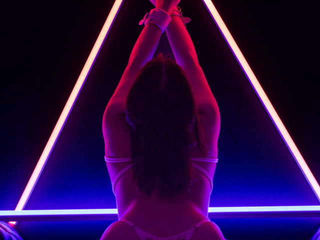 VickyButerfly with arms tied above her head, back facing camera, wearing white bra and thong, neon pink and purple triangle lights, dark background, wrists cuffed.