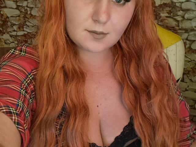 Kirobra69 with red wavy hair, plaid shirt, and black lace bra showing her big boobs. She has pale skin, dark lipstick, and a neutral expression.
