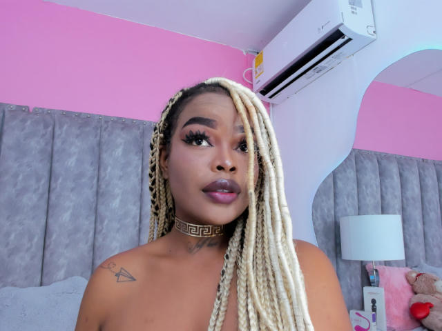 MadinsonLopez with blonde braids, dark skin, thick eyebrows, long eyelashes, glossy pink lips, gold choker, tattoo on left shoulder, looking to the side, air conditioning unit above.
