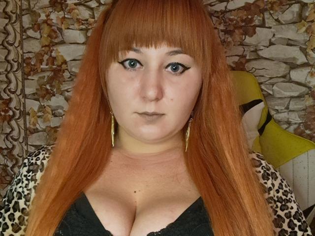 Kirobra69 has red hair, blue eyes, and is looking at the camera with a serious expression. She's wearing a leopard print top with black lace, and gold earrings.