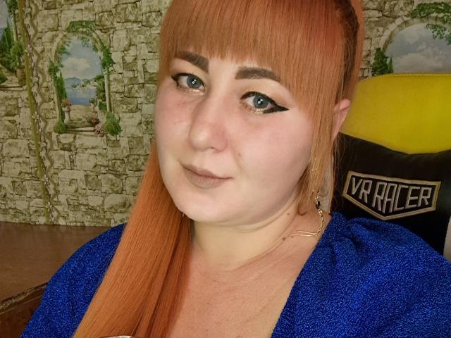 Kirobra69 with red hair, blue eyes, and black eyeliner, wearing a blue glittery top, smiling slightly. She has fair skin and a subtle nose. Her tits are small.