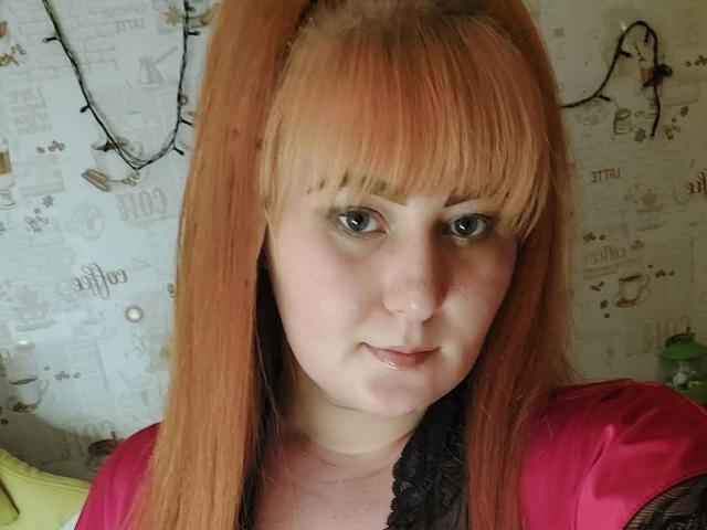 Kirobra69 has orange hair, blue eyes, and a slight smile. She's wearing a pink shirt with black lace. Her tits are small, and she has a cute nose.