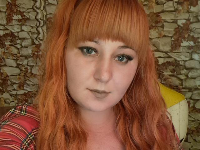 Kirobra69 has red, wavy hair with bangs, blue eyes, and gray lipstick. She's wearing a red plaid shirt, looking straight at the camera with a neutral expression.