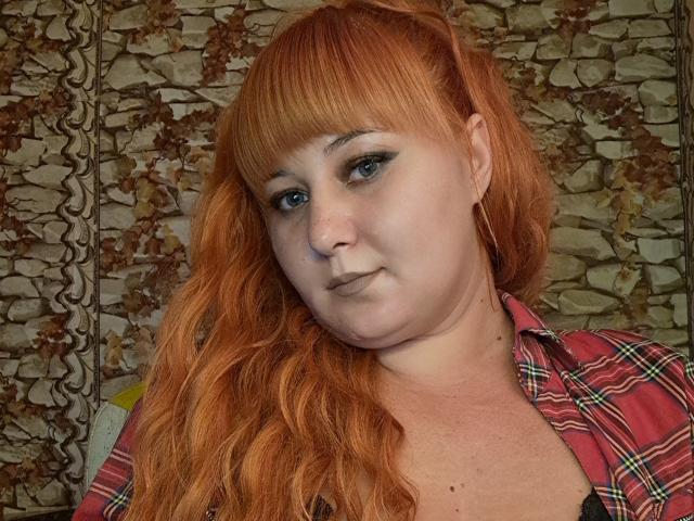 Kirobra69 has red hair, blue eyes, and is wearing a plaid shirt. Her tits are small and her face has a neutral expression. She's looking at the camera.