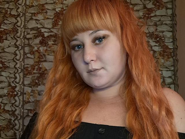 Kirobra69 has pale skin, orange wavy hair with bangs, blue eyes, and a slight pout. She's wearing a black top, looking at the camera.
