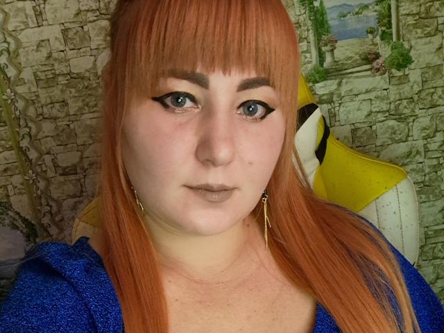 Kirobra69 has pale skin, orange hair, black eyeliner, and blue eyes, wearing a blue shirt with gold earrings, looking straight at the camera with a neutral expression.
