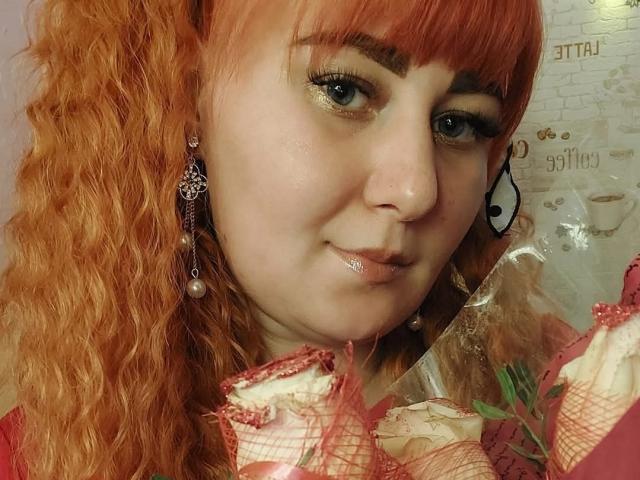 Kirobra69 with red curly hair, shiny gold eyeshadow, and pearl earrings, smiling at the camera, holding a red and white checkered fabric with lace edges.