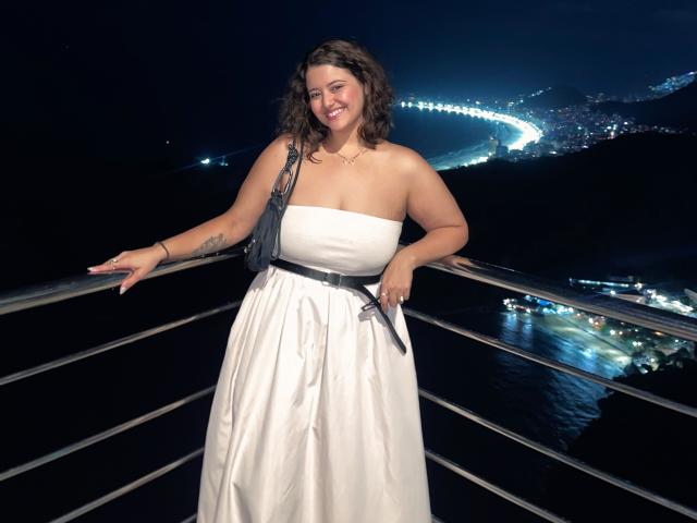 AgathaVelline with curly brown hair, smiling, wearing a white strapless dress, black belt, and black fringe bag, standing on a metal balcony at night, holding a black phone.