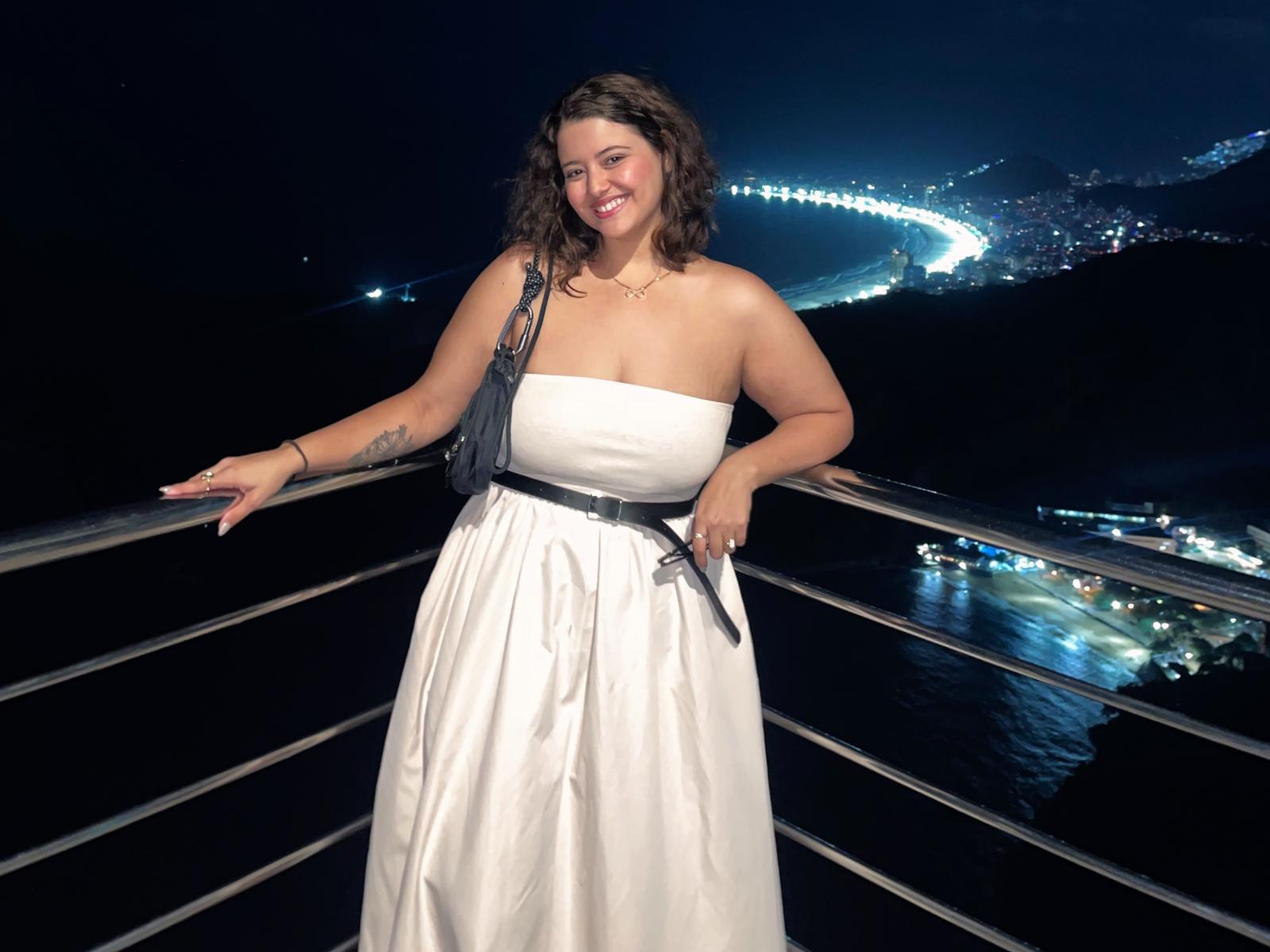 AgathaVelline with curly brown hair, smiling, wearing a white strapless dress, black belt, and black fringe bag, standing on a metal balcony at night, holding a black phone.