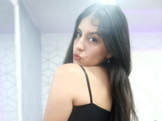 MadissonFontaine with long black hair, looking back over her shoulder, pouting, wearing a black spaghetti-strap top, showing her bare shoulder and small tits. Soft light, white background.