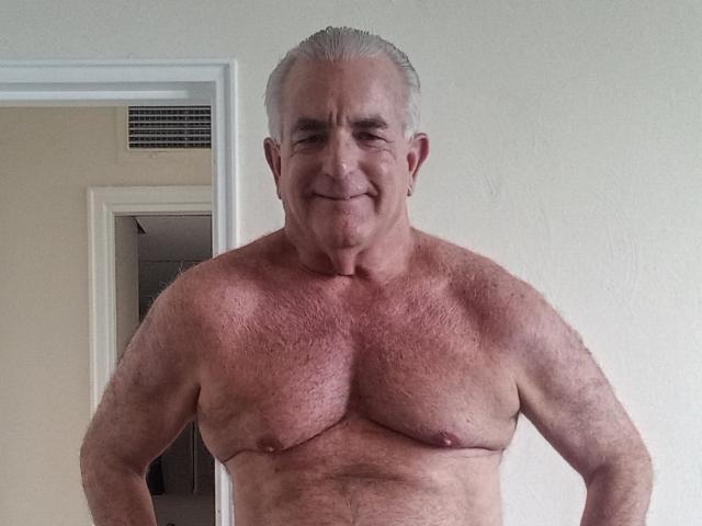 ShoreCock, an older, muscular man with gray hair, shirtless, showing off his hairy, large chest and a slight smile. His nipples are visible. He's standing with hands on hips.
