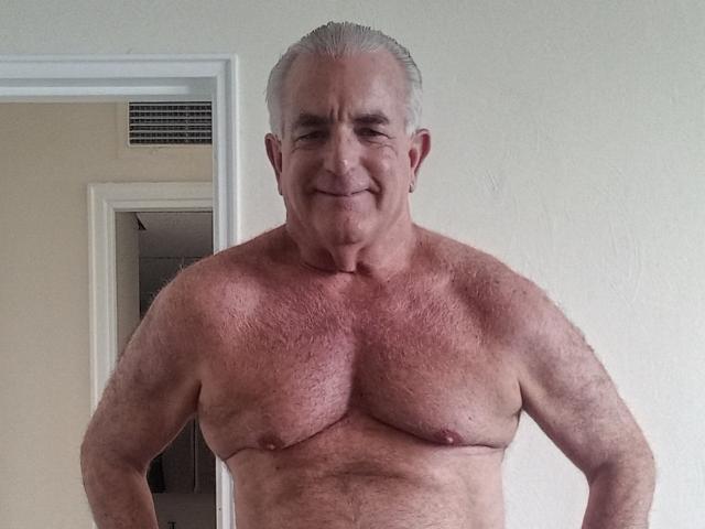 ShoreCock is an older, shirtless man with gray hair, a hairy chest, and a slight smile, standing in a doorway, showing his small, sagging, bare-assed, flabby tits.