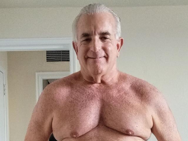 ShoreCock is an older, shirtless dude with gray hair, small tits, and a slight smile, standing in a room with a door and air vent in the background.