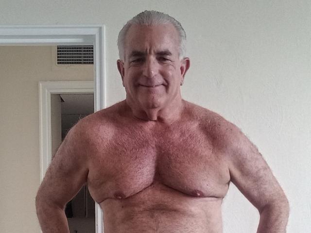 ShoreCock is an older, shirtless dude with gray hair, a slight smile, and a hairy, muscular chest. His small nipples are visible. He stands in a doorway.
