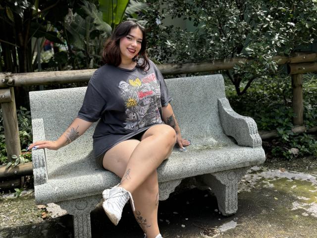 Milly Johnson, sentada en un banco de piedra, vestida con una camiseta gris estampada, pantalones cortos negros y zapatillas blancas, mostrando tatuajes en los brazos y las piernas, sonriendo con lápiz labial rojo, cabello oscuro..