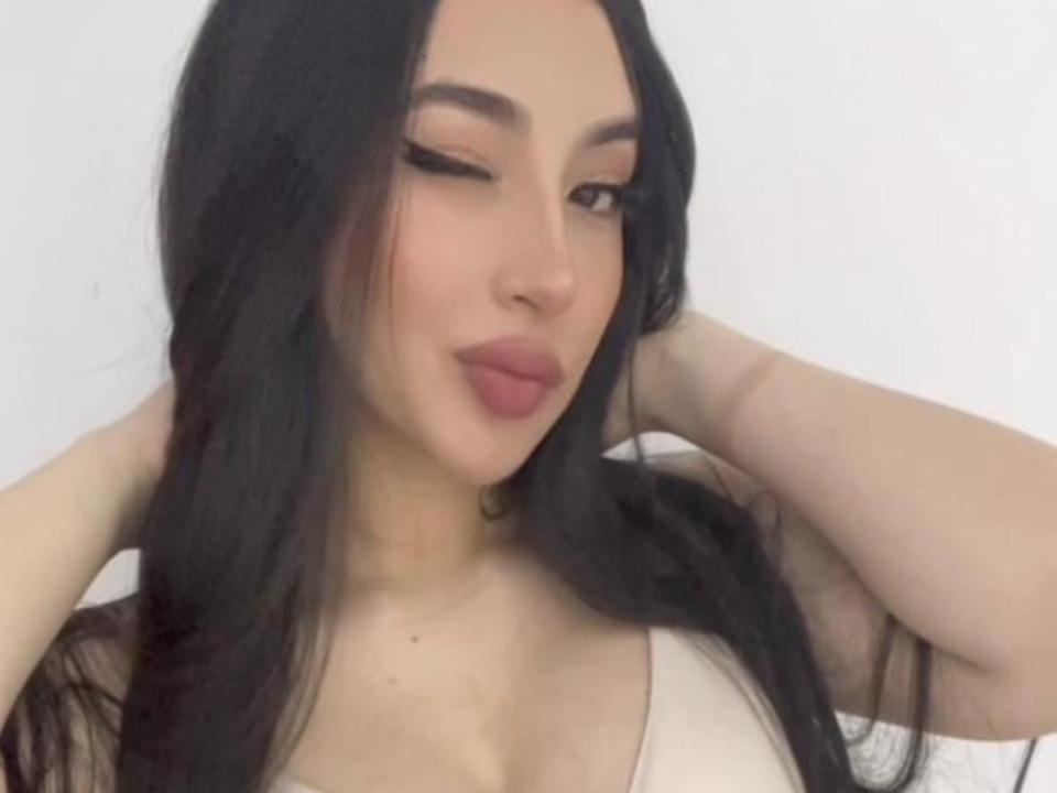 MiladyCute with long black hair, winking, hands in hair, wearing a white top, showing her boobs, pale skin, full pink lips, and subtle makeup.