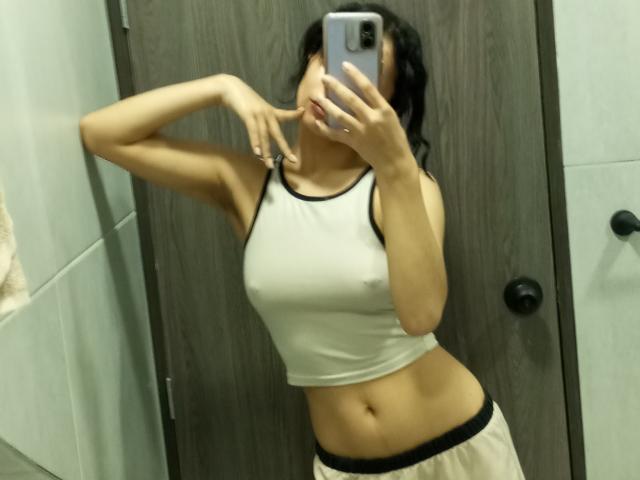 Heily Peterson, con i capelli neri, indossa una canottiera bianca che mette in mostra il seno e pantaloni beige, sta scattando un selfie allo specchio, con un dito sul mento, tenendo un telefono..