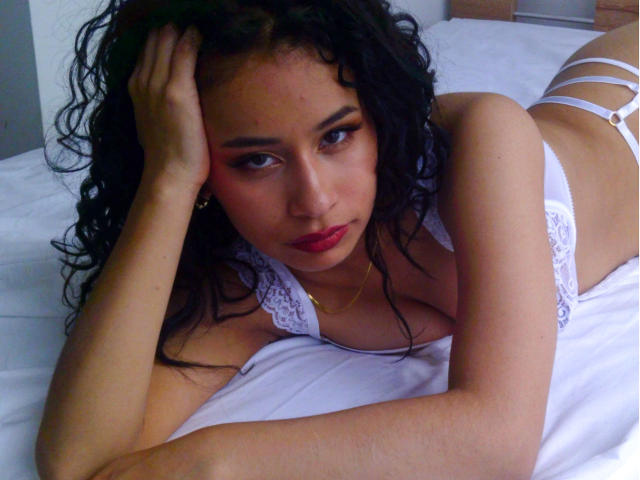 Heily Peterson, con i capelli neri ricci, sdraiata su un letto bianco, indossa un reggiseno bianco in pizzo e biancheria intima bianca con spalline, una mano sulla testa, guarda la fotocamera con rossetto rosso e eyeliner..