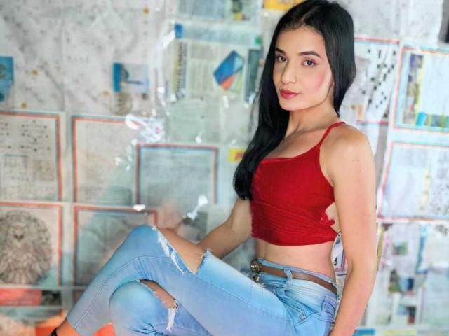 PamelaKander with long black hair, red tank top, and ripped blue jeans, sitting, showing a cute face, small tits, and a slim, sexy build.