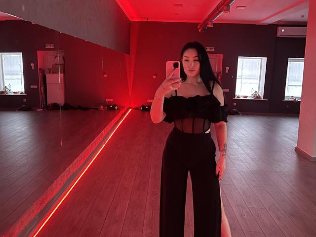 WinterJamie with black off-shoulder top showing tits, sheer black pants, holding phone, red-lit room, long black hair, tattoo on right arm, standing, mirror reflection.