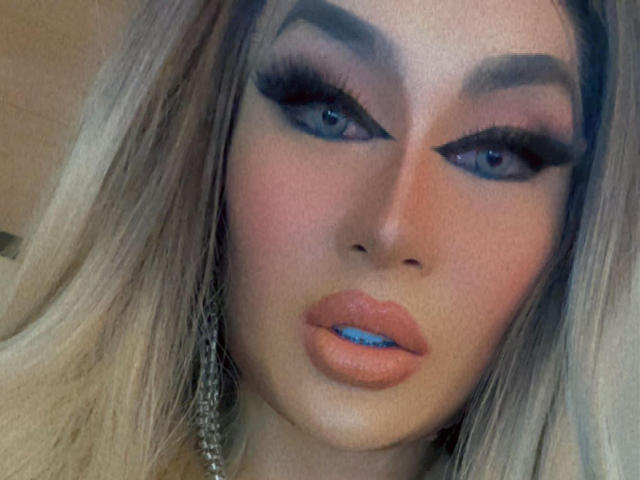 YourMistressoFevilx has blue lips, heavy black eyeliner, and long blonde hair with thick eyebrows. She's looking at the camera with a serious expression. Her makeup is bold.