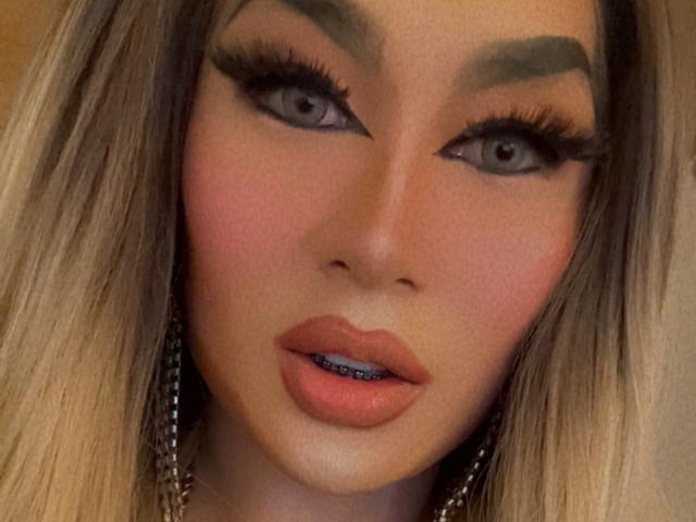 YourMistressoFevilx has blue eyes, thick black eyebrows, long eyelashes, pink blush, and pink lips with a slight open mouth showing a hint of blue lipstick.