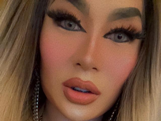 YourMistressoFevilx's face with bold eyebrows, grey eyes, thick eyelashes, pink blush, full lips with blue lipstick, and long blonde hair. Her expression is slightly open-mouthed.