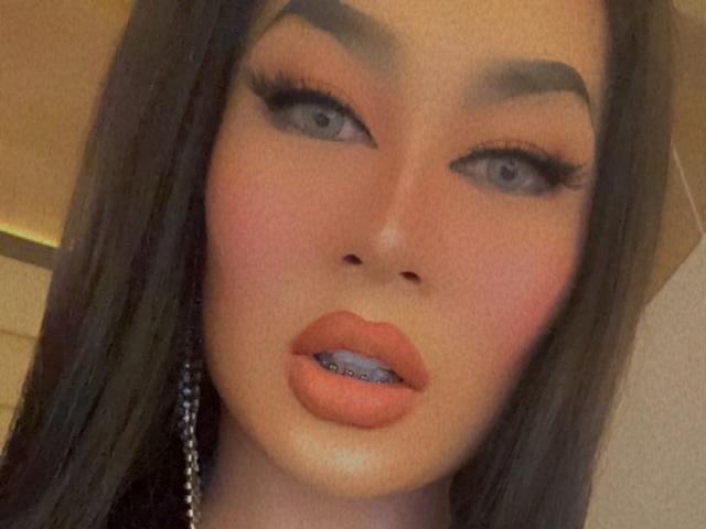 YourMistressofEvilx has full, glossy orange lips with a blue tooth, thick black eyeliner, and long, dark brown hair. Her eyebrows are thick and she's looking slightly to the side.
