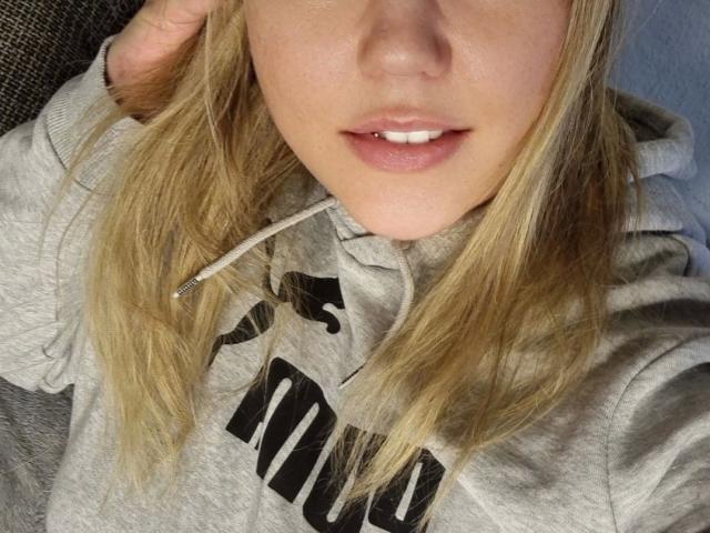 LiesaBDSM-hot's face, blonde hair, slightly parted pink lips, grey hoodie, visible white teeth, right hand in hair, soft light, casual, relaxed, upper torso, close-up.