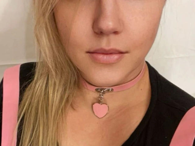 LiesaBDSM-hot's face, showing pink choker with heart pendant, black shirt, pink straps, blonde hair, light skin, and pink lips. Her tits are not visible.