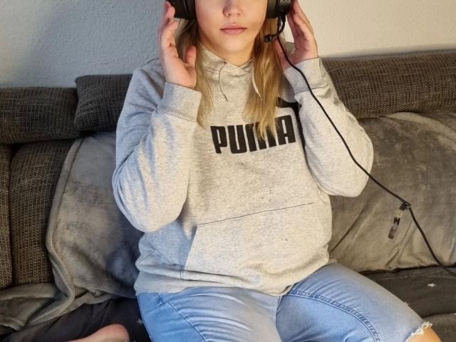 LiesaBDSM-hot with headphones on, grey hoodie, and ripped jeans, sitting on a couch, hands on headphones, blonde hair, light skin, subtle smile.