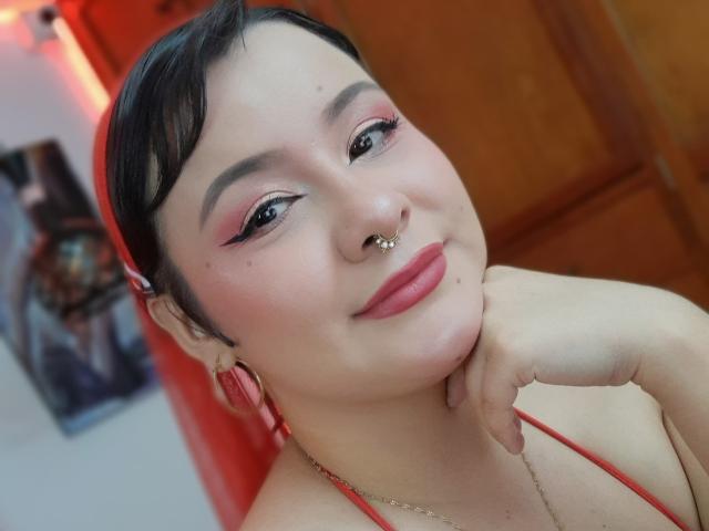 CassandraSexy with short black hair, pink eyeshadow, black eyeliner, nose ring, septum ring, pink lipstick, red earrings, red halter top, and hand on cheek, smiling.