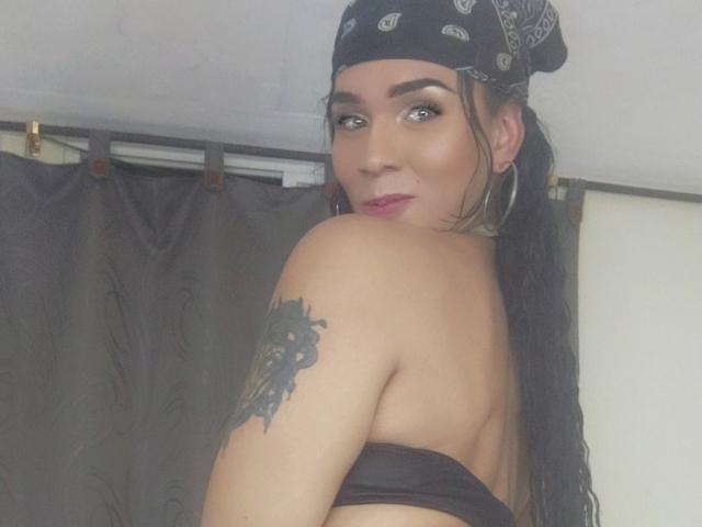 DiosaV with long black hair in a bandana, hoop earrings, and black strapless top, showing tattooed shoulder, looking back with pink lipstick, slightly smiling.