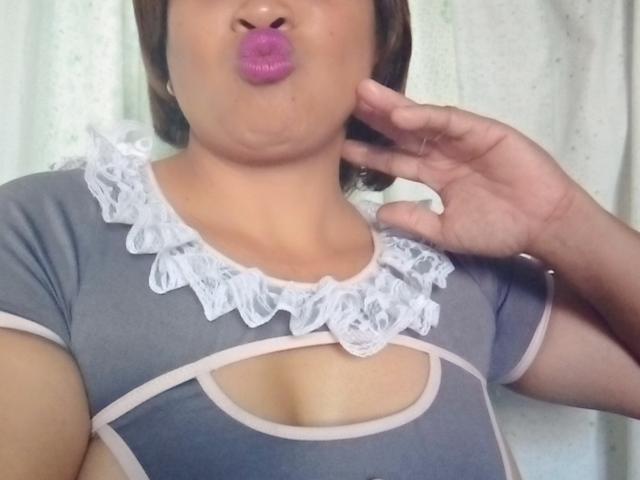Rahel69 avec du rouge à lèvres rose, une robe grise avec des détails en dentelle blanche, et une découpe laissant entrevoir ses seins, des joues gonflées, une main touchant sa joue, des cheveux courts et bruns, une peau claire..