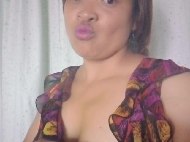 Rahel69 with puffy pink lips, brown skin, and a colorful ruffled top, showing her cleavage and small to medium-sized tits. She's making a pouty face.