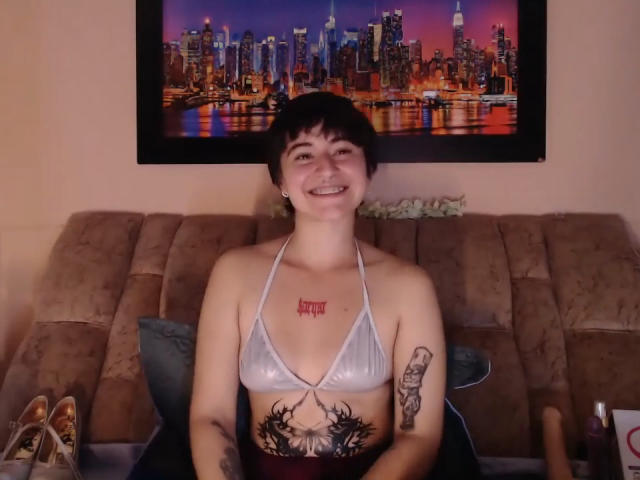 Short-haired MissTat with small tits, wearing a silver bra, sitting on a brown couch, smiling, showing tattoos on chest and arm, city skyline wall art behind.