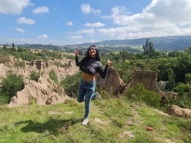 Xewaa with big tits, wearing a black crop top and blue jeans, smiling and flipping double peace signs, standing on grassy terrain with rocky formations and a green landscape.