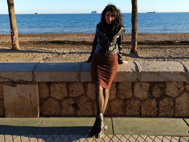 AddictedToUTS, com cabelo escuro e encaracolado, jaqueta de couro preta, saia vermelha texturizada e botas cinzas, apoiada em uma parede de pedra, em um caminho de azulejos, perto de um mar azul..