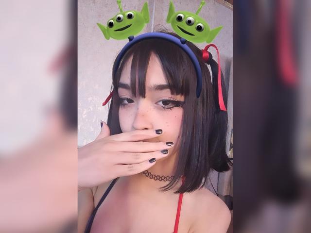 MillaHottLove69 with black bob, green alien headbands, red ribbon, choker, black nail polish, hand covering mouth, looking at mirror, soft light, gentle expression.