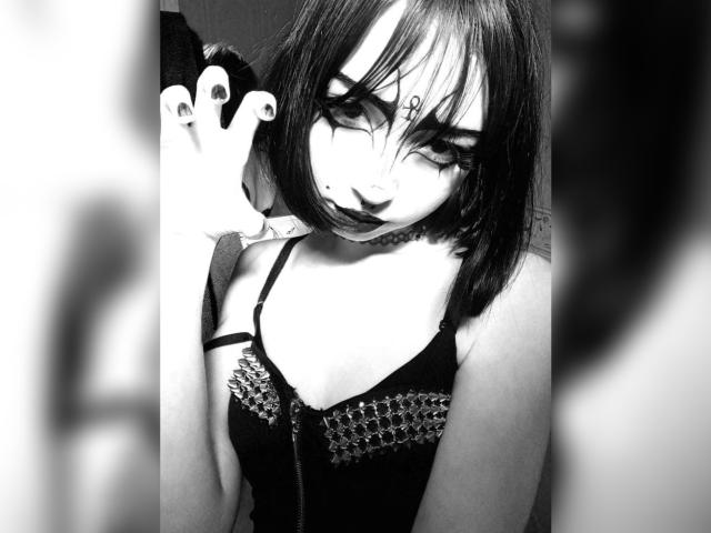 Black-and-white photo of MillaHottLove69 with short dark hair, goth makeup, black lipstick, eyeliner, wearing a spiked dress, showing a finger with painted nails, looking seductively.