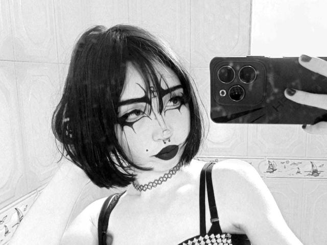 Black-and-white selfie of MillaHottLove69 with short dark hair, goth makeup, black lipstick, nose ring, choker, and striped top, holding a camera, showing her tits.