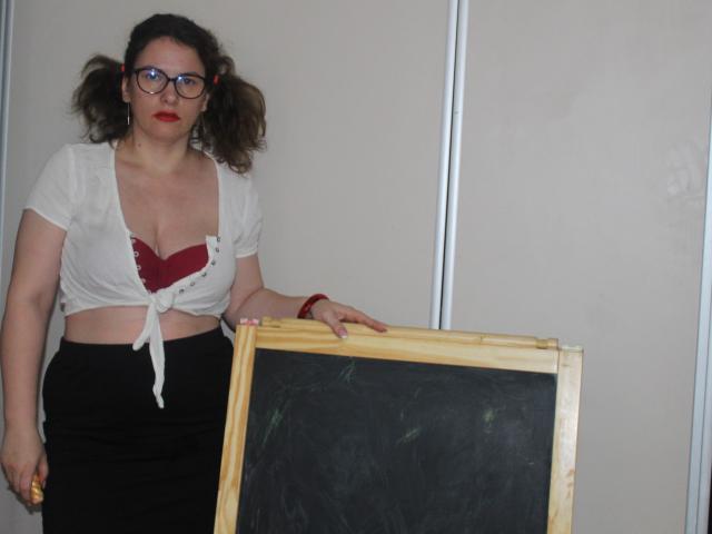 KinkyKosmina with curly brown hair, glasses, red lipstick, white tied-up top showing red bra, black skirt, holding chalkboard, standing, right hand on chalkboard, left hand down.