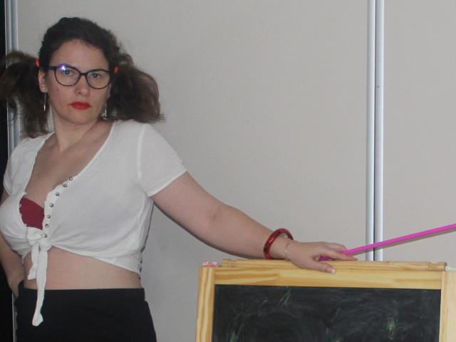 KinkyKosmina with puffy brown hair in pigtails, wearing glasses, red lipstick, white tied-up shirt showing red bra, black skirt, holding pink pointer, standing by chalkboard.